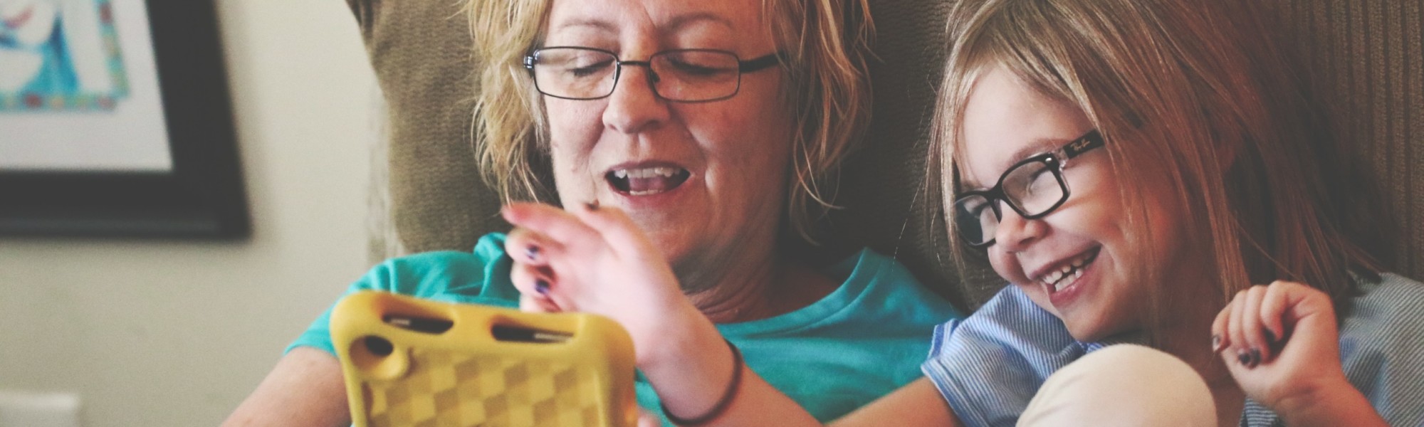 Isoäiti ja lapsi nauravat sohvalla, selaavat tablettia
