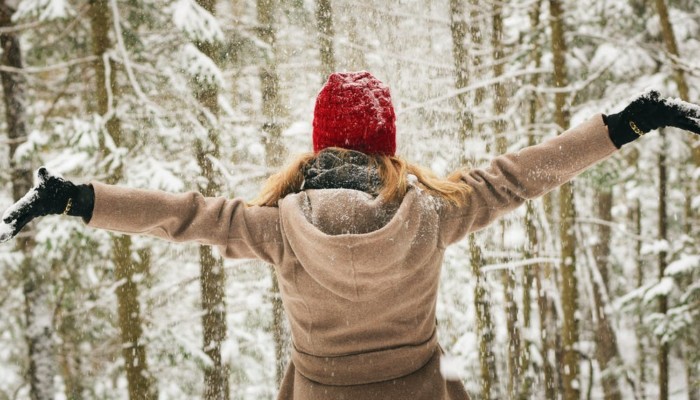 Nainen kädet levällään lumisessa metsässä, selkäpuolelta kuvattuna.