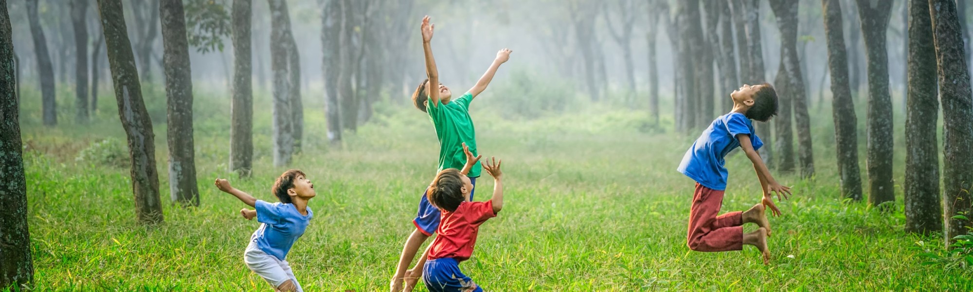 Kuvassa on neljä ilmaan hyppäävää iloista lasta vihreän luonnon keskellä.
