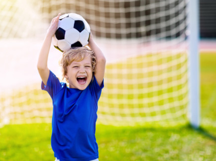 Iloinen poika pitää jalkapalloa päänsä yläpolella, takana näkyy jalkapallomaali