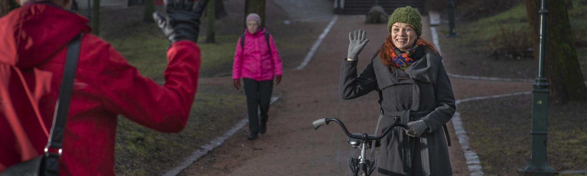 Nainen taluttaa pyörää ja tervehtii vastaantulijaa kädellä