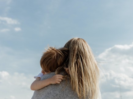 Kuvassa pitkähiuksinen nainen pitää sylissään lasta, joka halaa häntä tiukasti. Nainen on kuvattu selkäpuolelta ja taustalla näkyy sininen taivas.