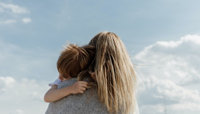 Kuvassa pitkähiuksinen nainen pitää sylissään lasta, joka halaa häntä tiukasti. Nainen on kuvattu selkäpuolelta ja taustalla näkyy sininen taivas.