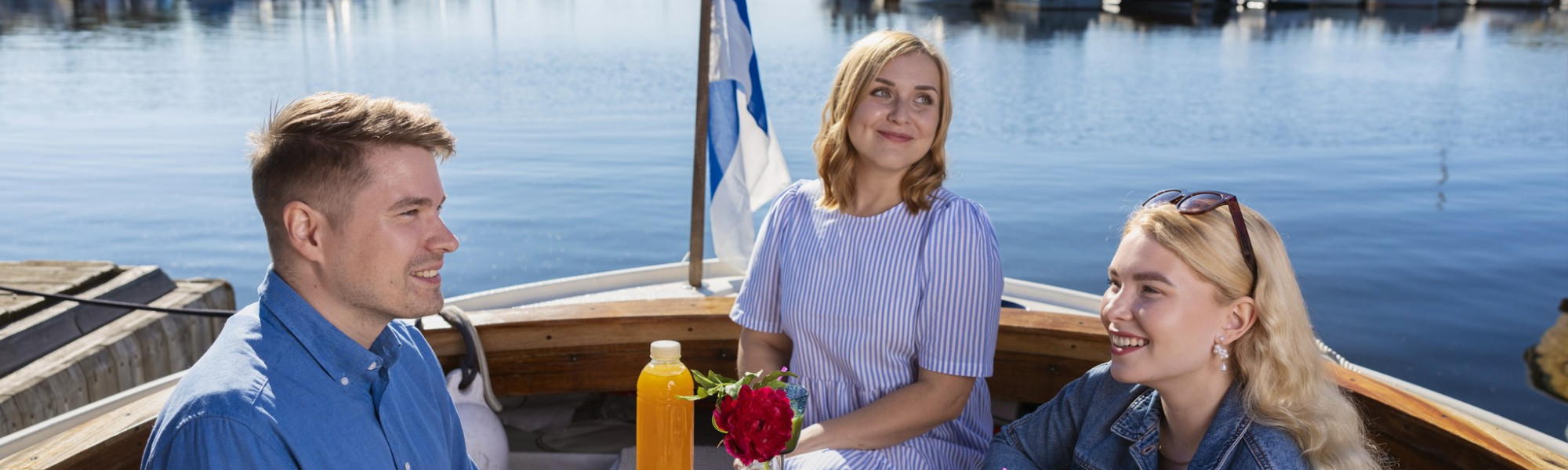 Kuvassa ihmiset syövät eväitä veneellä. Taustalla näkyy vesistö ja Suomen lippu.