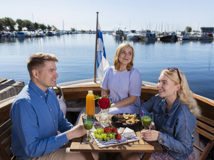 Kuvassa ihmiset syövät eväitä veneellä. Taustalla näkyy vesistö ja Suomen lippu.