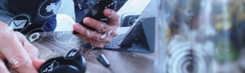 Käsi käyttää VOIP-kuulokemikrofonia sekä näppäimistöön telakoitua tablettia ja älypuhelinta. Konsepti liittyy viestintään, IT-tukeen, puhelinpalveluun ja asiakaspalveluun. Etualalla maljakko kukka, näytöllä virtuaalisia käyttöliittymäkuvakkeita.