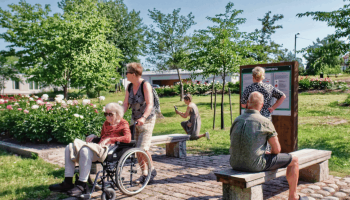 Ihmisiä kesäisessä puistossa. Etualalla pyörätuolissa iäkäs rouva.