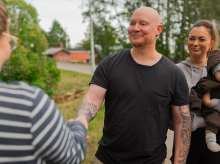 Mies kättelee kaupanteon merkiksi. Nainen vauva sylissä hymyilee hänen takana.