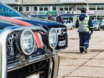 Etualalla autojen keuloja, taustalla pysäköinninvalvojia kävelemässä poispäin.