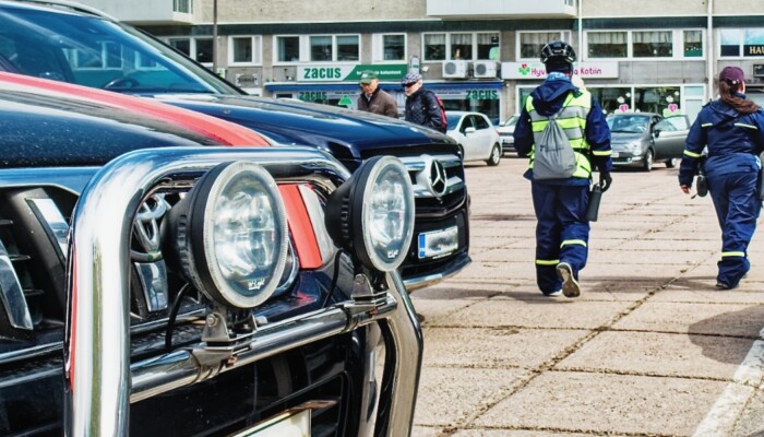 Etualalla autojen keuloja, taustalla pysäköinninvalvojia kävelemässä poispäin.
