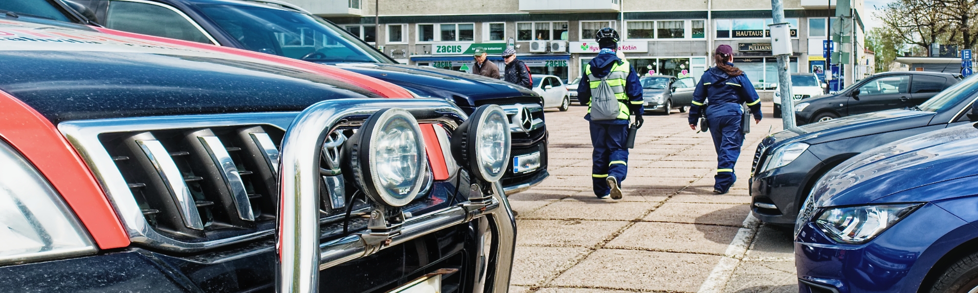 Etualalla autojen keuloja, taustalla pysäköinninvalvojia kävelemässä poispäin.
